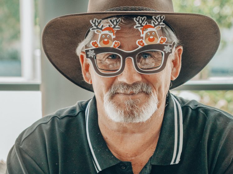 Image of An older man with a white beard wears a brown wide-brimmed hat and fun glasses decorated with cartoon reindeer. He is dressed in a black polo shirt, and is smiling slightly, sitting indoors with a blurred background.