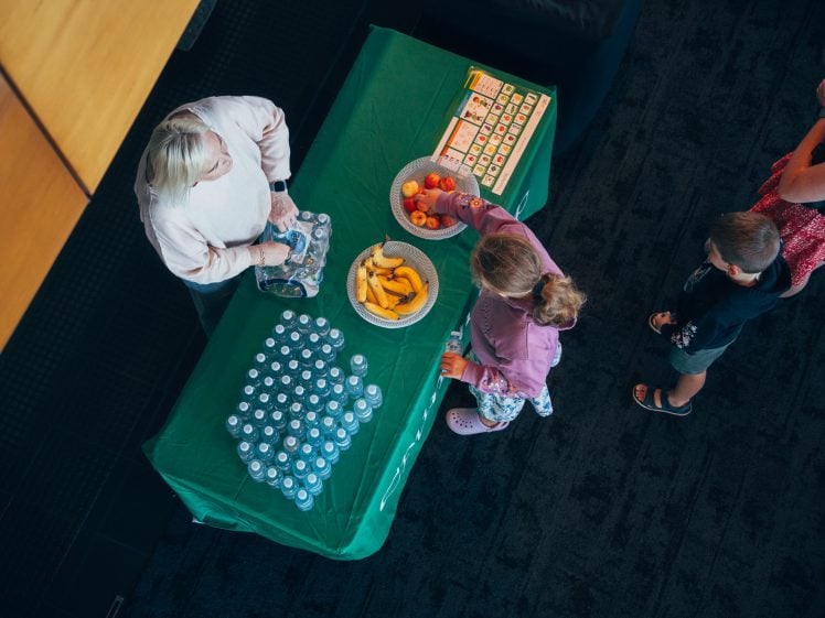 Image of A woman stands behind a green-covered table with water bottles, bananas, apples, and snacks, while a child reaches for fruit and two other children stand nearby. The scene is viewed from above.