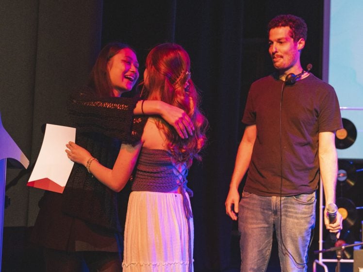 Image of Two women hug and smile on stage whilst a man stands nearby holding a microphone, looking at them. One woman holds a piece of paper. Stage equipment and a screen are visible in the background.