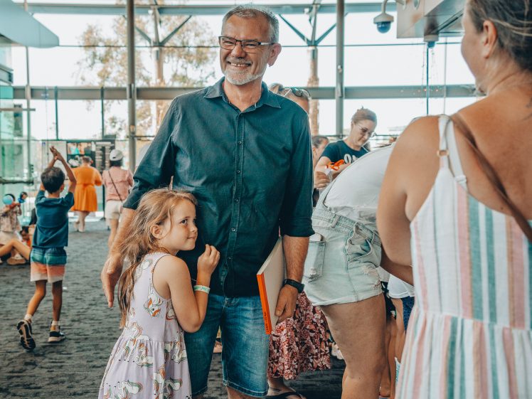 Image of A man smiles whilst holding a folder, with a young girl hugging him. They are indoors with several people, including children, visible in the background. The scene appears lively and sociable.