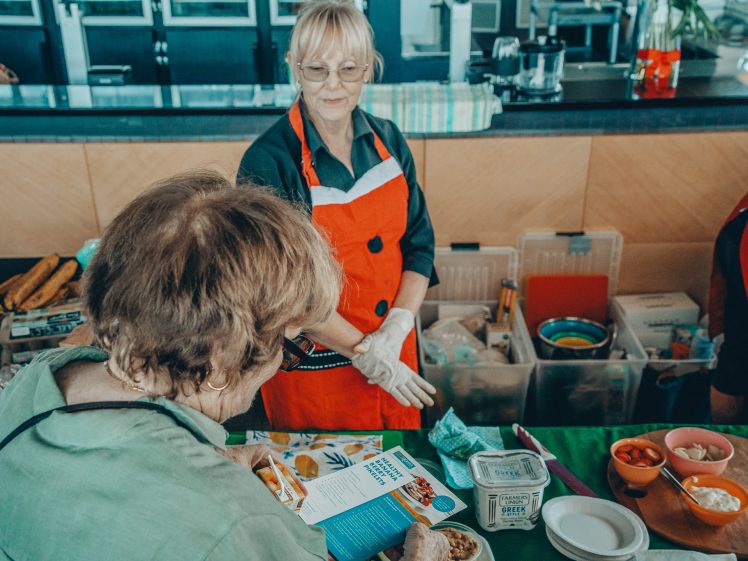 Image of Two women at a market or food stall; one in an orange apron and gloves stands behind the counter, while the other examines a leaflet. Various food items and containers are arranged on the table between them.