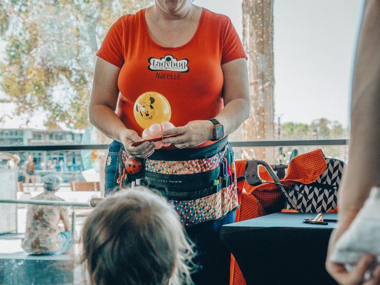 Image of A woman with glasses and a bright orange shirt makes a balloon animal for a young child indoors. She has small balloons on her head and a colourful pouch, whilst the child watches eagerly from the foreground.