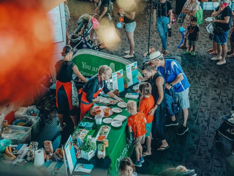 Image of A group of adults and children gather around a green stall at an indoor event, where staff in aprons serve food samples and share information. Other people and pushchairs are visible in the background.
