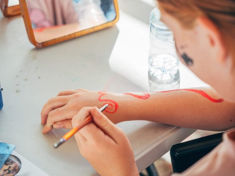 Image of A person uses a brush to paint a red swirl on their forearm whilst sitting at a table with art supplies, a mirror, and a water bottle.