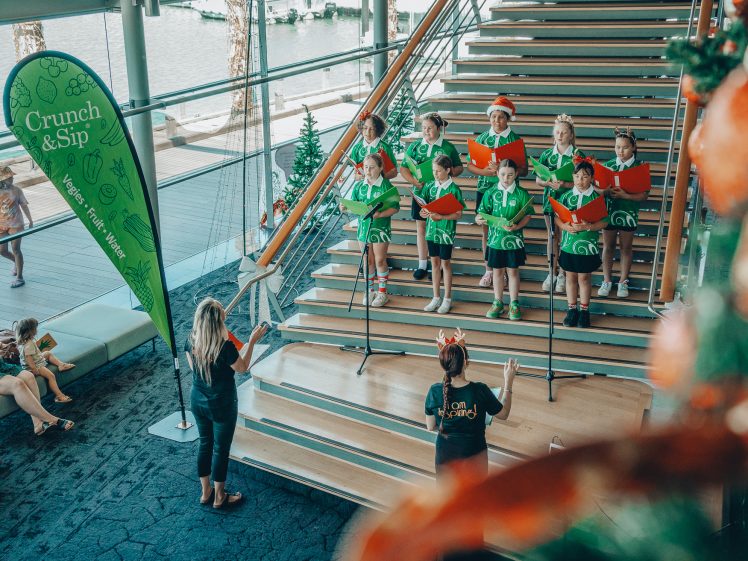 Image of A children’s choir in green shirts and Santa hats sings on wooden stairs, holding red songbooks, while two adults conduct. Onlookers sit nearby. A “Crunch & Sip” banner and holiday decorations are visible.