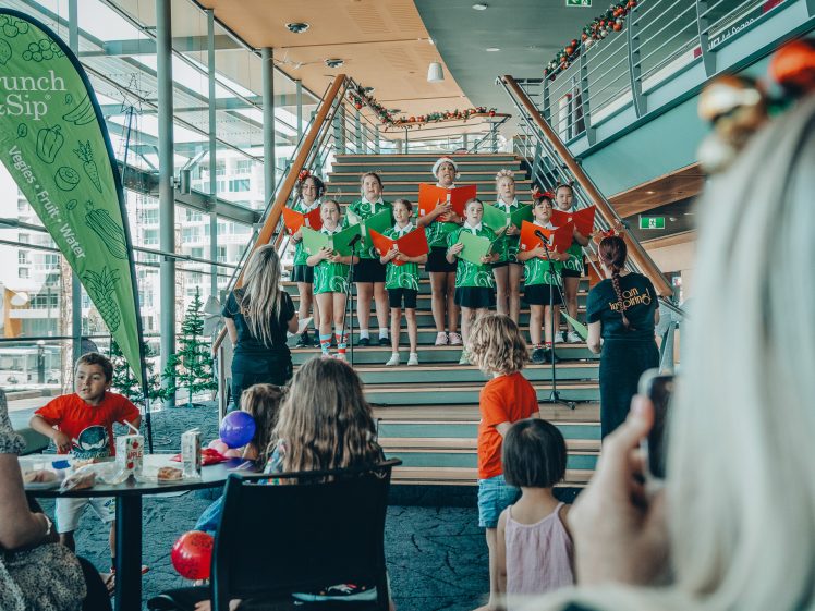 Image of A group of children stand on indoor stairs singing in a choir, holding green and red songbooks. Audience members, including children and adults, watch from nearby tables. Festive decorations and a Crunch & Sip banner are visible.