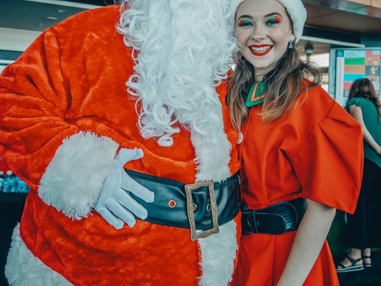 Image of A person dressed as Father Christmas stands next to a smiling woman in a red and green elf costume and Father Christmas hat. They are indoors, posing together for a holiday-themed photo.