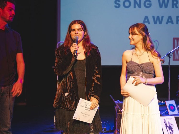 Image of Three people stand on stage at an awards event. The person in the centre speaks into a microphone, holding a certificate. A woman in a white dress smiles at them. A large projection screen with award text is visible behind them.