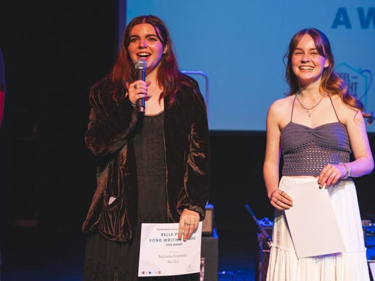Image of Two young women stand on stage smiling, one holding a microphone and an award certificate, the other holding a piece of paper. They are lit by stage lights, with a blue award presentation screen behind them.