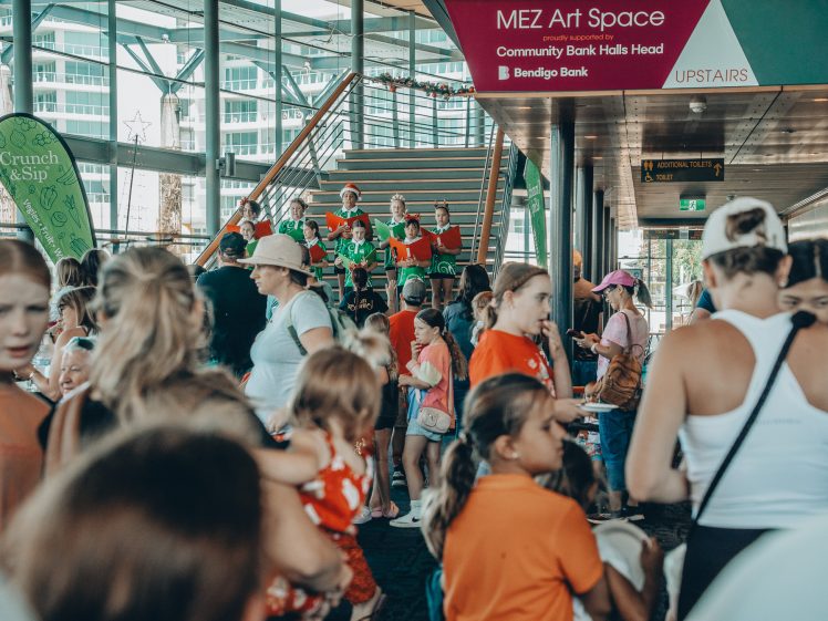 Image of A large group of adults and children gather indoors at a busy event. Some children in green and red shirts sit on stairs in the background, whilst others interact and play throughout the crowded space. Signs point to an upstairs art space.