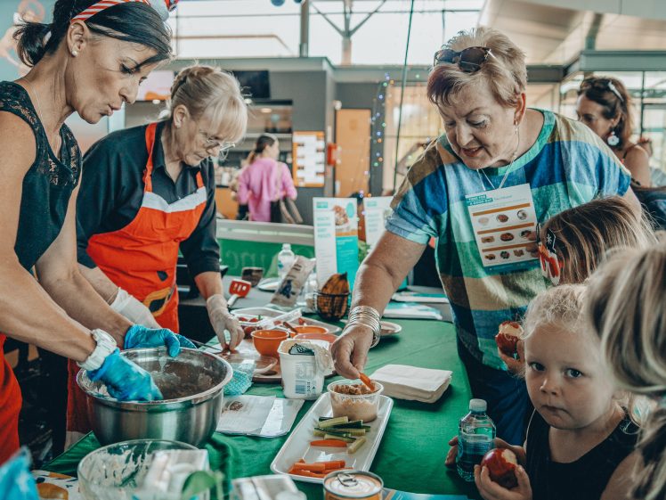 Image of A group of adults and children gather around a table with food samples at an indoor event. Two women prepare food whilst others look on. A young child in the foreground holds a snack and looks at the camera.