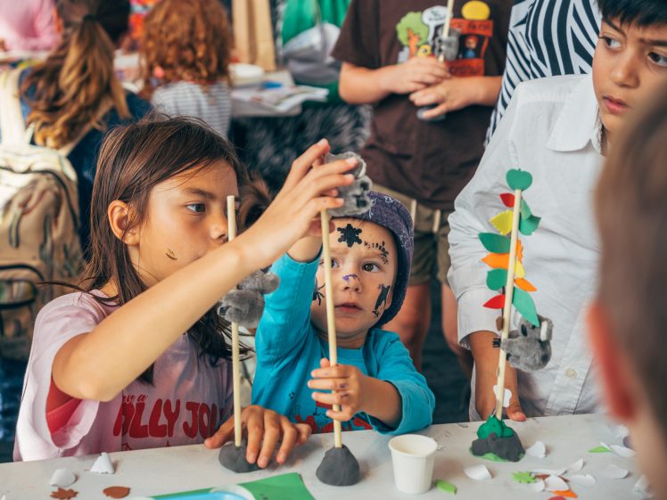 Image of Children sit at a table engaged in a craft activity, building structures with clay, sticks, and paper shapes. One child has face paint and reaches up to adjust an object, whilst others watch and participate.