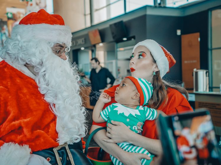 Image of A woman in a Santa hat holds a baby dressed as an elf, both looking at a man dressed as Father Christmas. The scene appears festive, indoors, with another person and a mobile in the background.