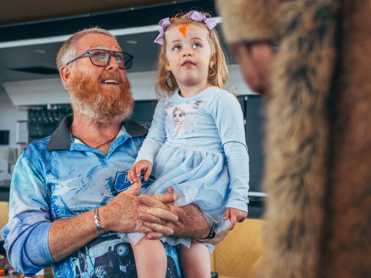 Image of A bearded man wearing glasses holds a young girl dressed in a blue dress and flower headband. They both look up, smiling, at a person in a furry costume indoors.