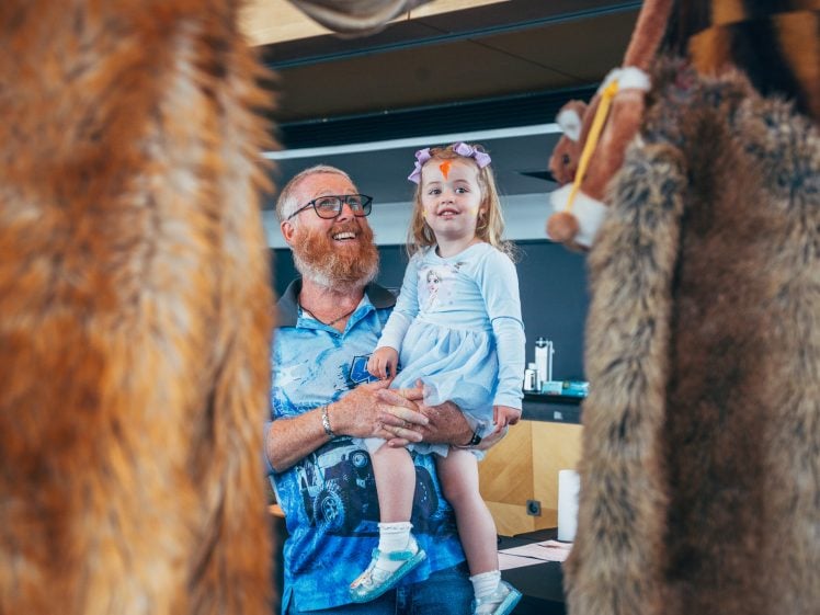 Image of A smiling man holding a young girl in a blue dress, both looking at something off-camera, framed by two large, furry animal costumes in the foreground.