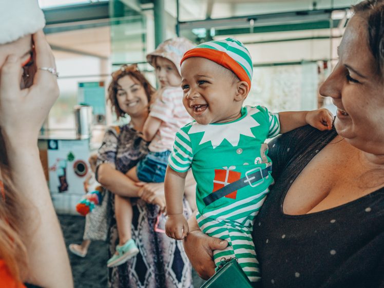 Image of A smiling baby in a green elf costume is held by a woman, surrounded by joyful adults and another baby in a festive indoor setting.