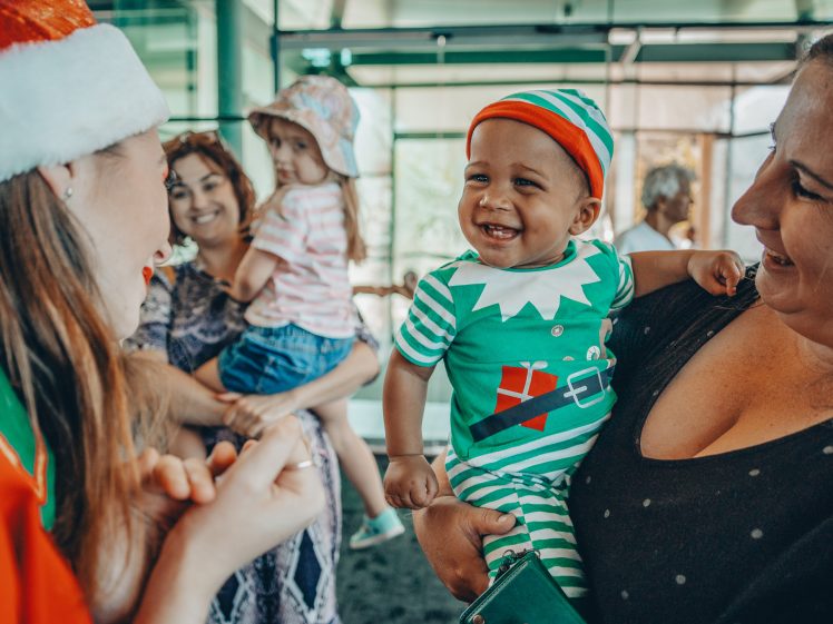 Image of A smiling baby in a green elf costume is held by a woman, while another woman in a Santa hat interacts with them. In the background, a woman holds another child. Everyone appears happy and festive indoors.