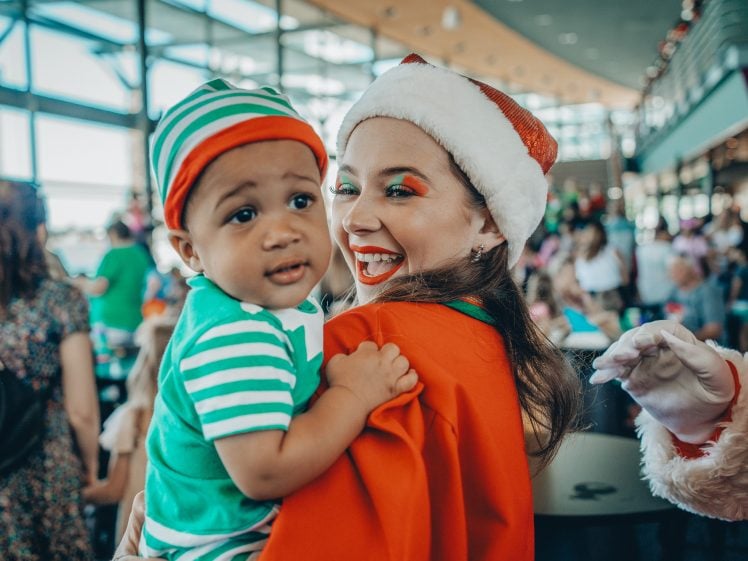 Image of A smiling woman in a Santa hat holds a baby dressed in green and white stripes at a festive indoor event, surrounded by people in the background.