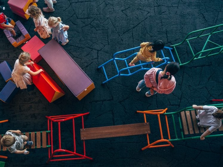 Image of Children play on colourful climbing frames and soft blocks arranged on a dark, textured floor. They are climbing, walking, and interacting in a busy indoor play area, viewed from above.