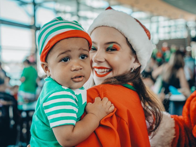 Image of A smiling woman in a Santa hat holds a baby dressed in a green and white striped outfit. They are indoors, and the background is filled with people and natural light from large windows.