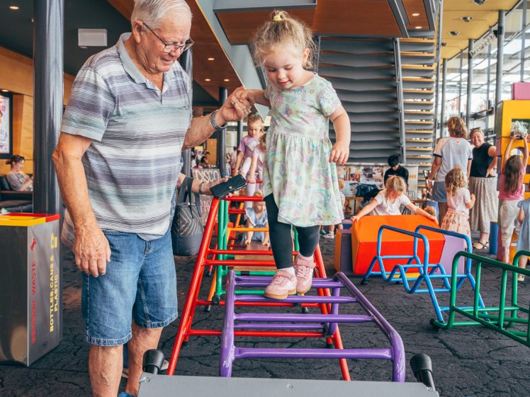 Image of An elderly man helps a young girl balance as she walks on colourful playground bars indoors, with other children and adults in the background.