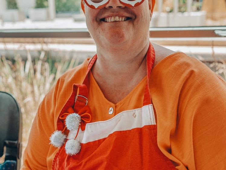 Image of A smiling person wearing a festive orange and red outfit, Santa-themed glasses, and a headband with decorations sits at a table with papers outdoors, celebrating in a cheerful holiday setting.