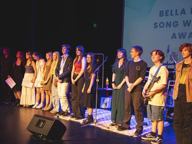 Image of A group of young people stand onstage in a line, smiling, at an awards event. Musical instruments and microphones are behind them. A screen displays “Bella Pe Song Writing Award”.
