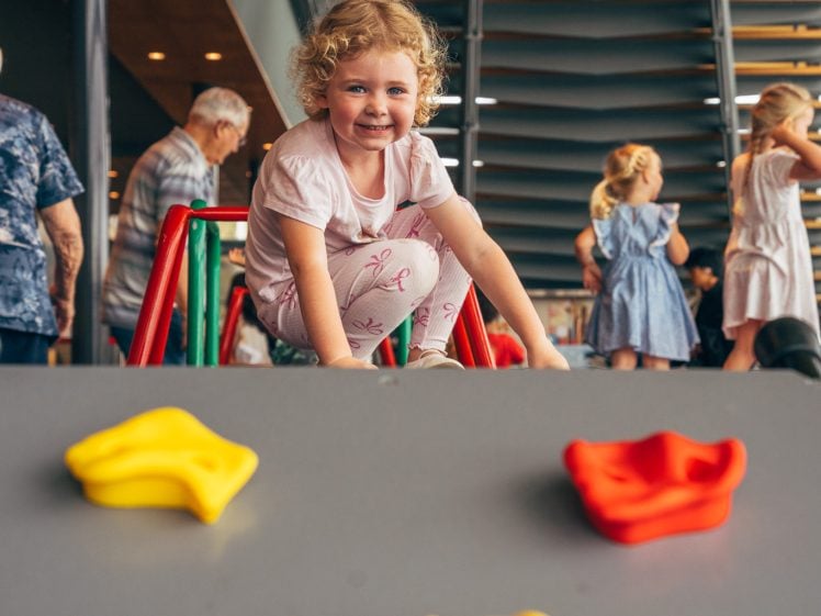 Image of A young child with curly hair and a pink outfit smiles while sitting at the top of an indoor playground slide. Colourful climbing holds are visible in the foreground, with other children and adults in the background.