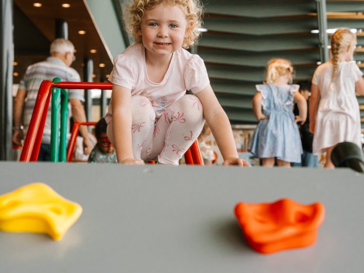 Image of A young child with curly blonde hair and a white shirt smiles while kneeling on playground equipment indoors. Other children and adults are visible in the background. Three colourful plastic shapes are in the foreground.