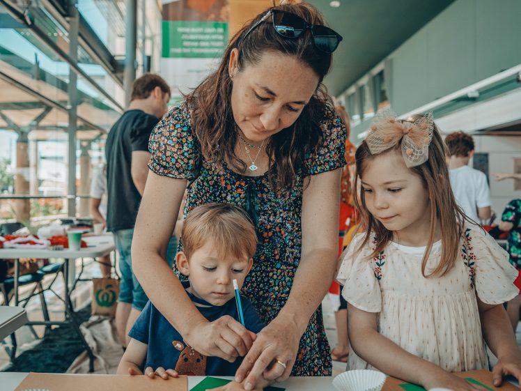 Image of A woman helps a young boy with a craft activity at a table, whilst a young girl stands beside them watching. They are indoors with other people in the background.