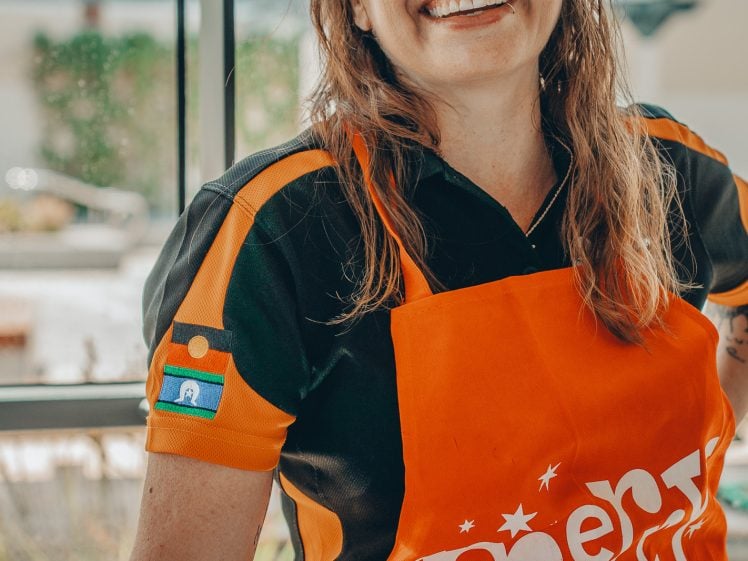Image of A smiling woman wearing festive green glasses and an orange apron that reads Merry Christmas stands indoors near a large window. She is dressed casually and looks cheerful.