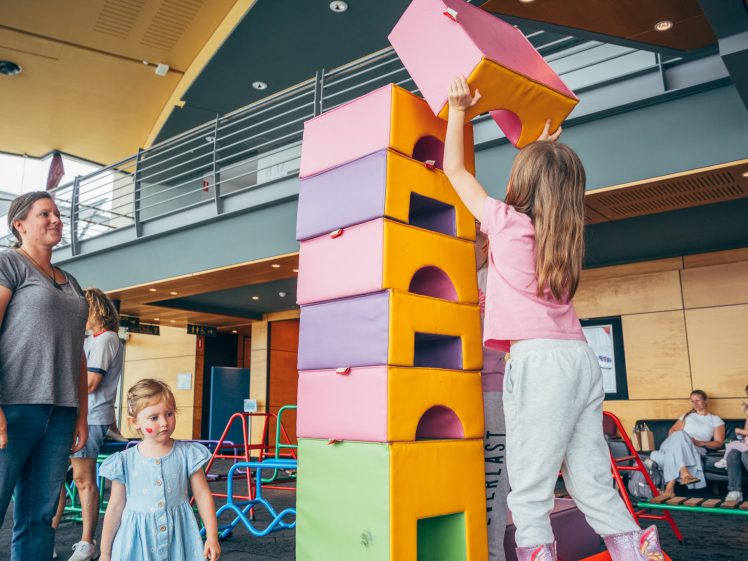 Image of A young girl stacks large, colourful blocks indoors while another child and an adult watch. People sit and watch in the background. The scene appears playful and lively.