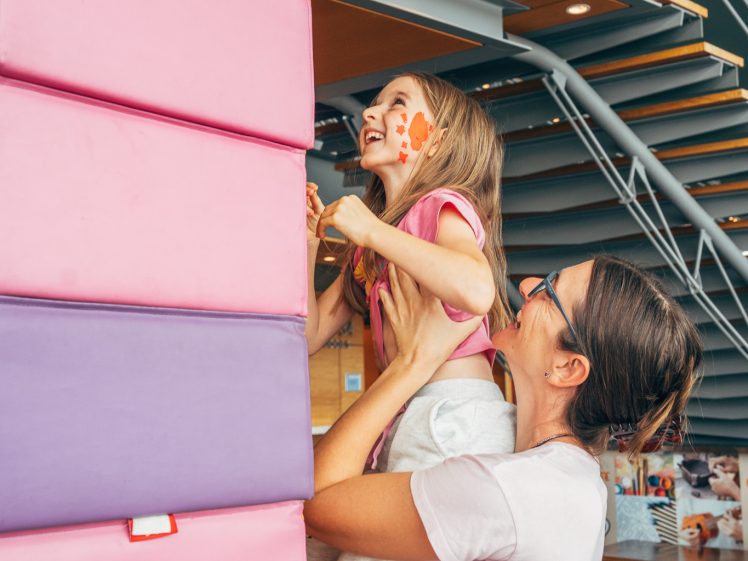 Image of A woman lifts a smiling child with face paint, helping her reach the top of a large stack of colourful foam blocks inside a play area, with a staircase in the background.