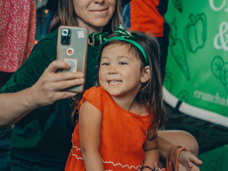 Image of A woman in a black hat takes a selfie with a smiling young girl in a red dress. They are at an event with people and a green, branded background behind them.