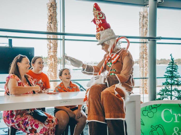 Image of A person in a whimsical gingerbread costume and tall hat sits by a table, playfully interacting with a smiling woman and two children. Christmas trees and festive décor are visible in the background.