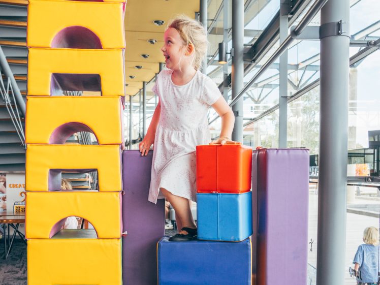 Image of A young girl in a white dress stands on large colourful foam blocks, smiling and playing inside a bright indoor play area with big windows and modern architecture.
