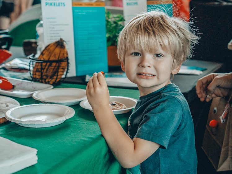 Image of A young boy with blond hair smiles at the camera while standing at a green table with food plates. Behind him, people are gathered near recipe posters and a display of fresh herbs at an indoor event.