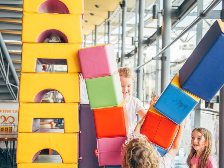 Image of Three young children play indoors, building a tall structure with large colourful foam blocks. Sunlight streams through large windows in the modern play area, creating a cheerful and lively atmosphere.