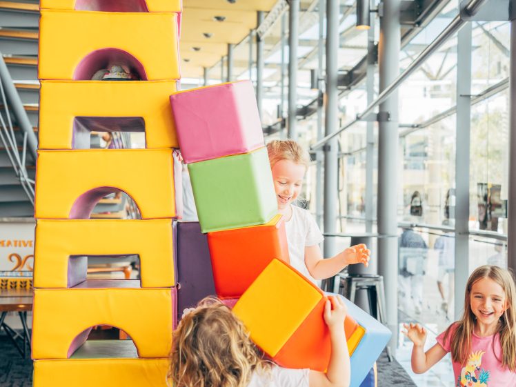 Image of Three young children play indoors with large colourful foam blocks, stacking them into a tall tower. The space is bright with natural light from large windows, and the kids are smiling and energetic.