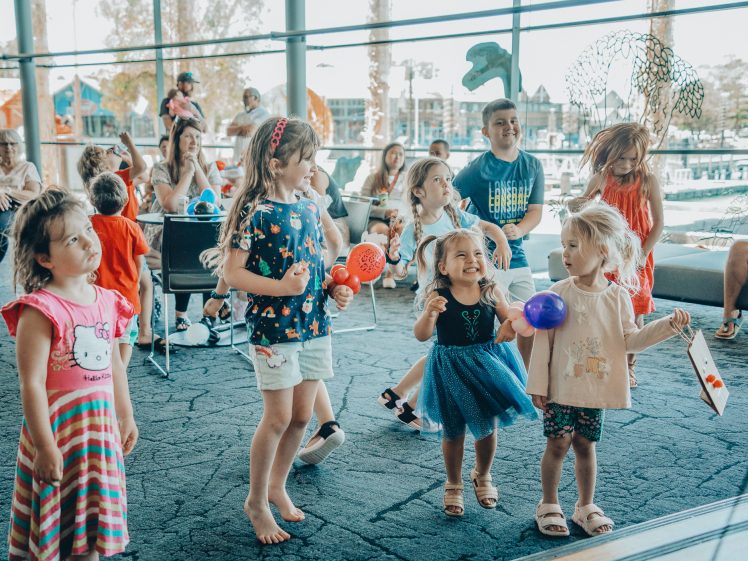 Image of A group of young children smile and play with balloons at an indoor event, whilst adults watch in the background. Large windows behind them reveal an outdoor scene with trees and water.