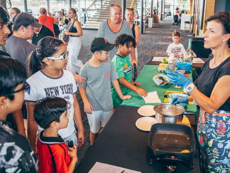 Image of A woman leads a cookery demonstration for a group of children and adults gathered around a table with cooking equipment in a bright, modern indoor space.
