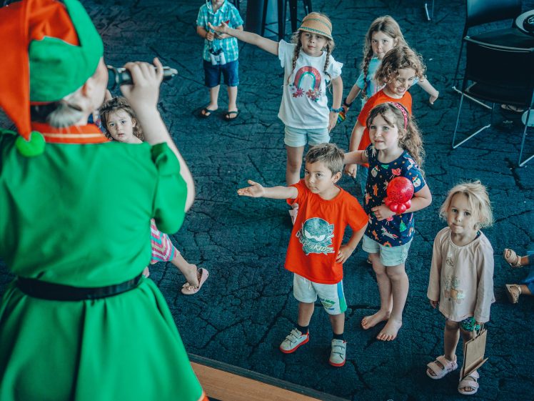 Image of A group of children stand in front of a performer dressed as an elf, some reaching out and others watching attentively in a brightly lit indoor space.