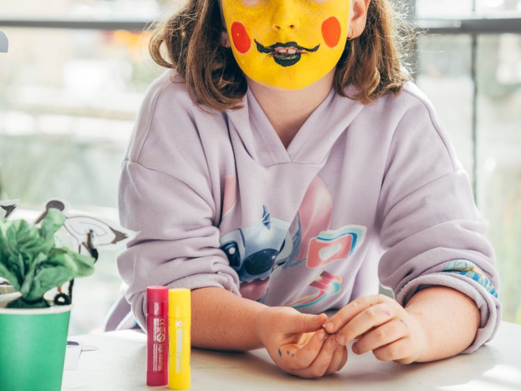 Image of A young girl with yellow face paint, red cheeks, and a black moustache and smile sits at a table, wearing a lavender hoodie with a cartoon design. She is indoors, with a plant and felt-tip pens on the table.