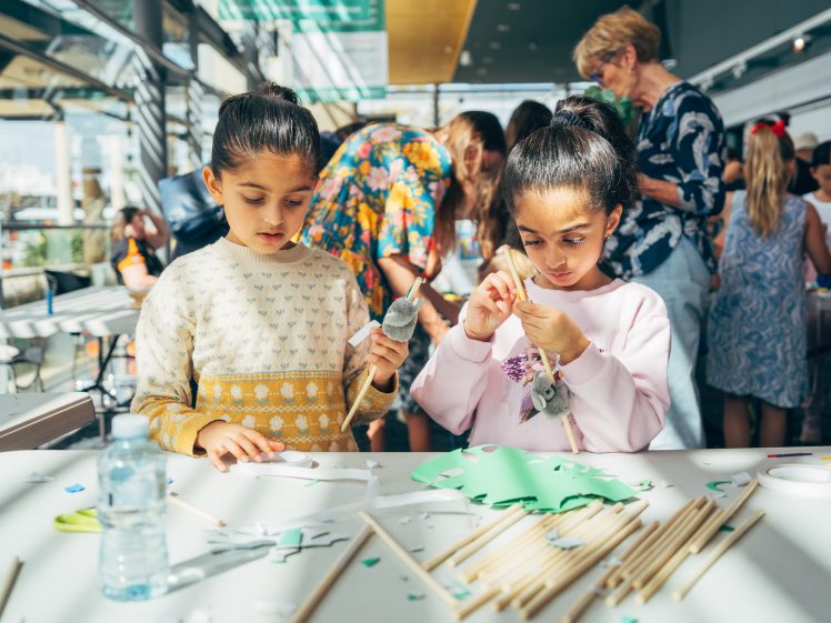 Image of Two young girls are focused on a craft project at a table covered with paper, sticks, and craft materials. Other children and adults are working in the background in a bright, busy indoor space.