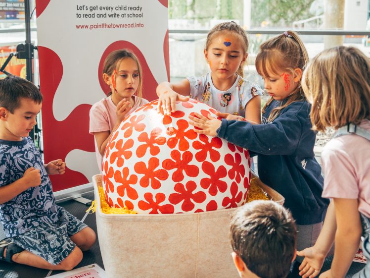 Image of A group of children decorate a large egg covered with red flower patterns at an indoor event. They are surrounded by art supplies and a banner promoting child literacy, with the slogan Let's get every child ready to read and write at school.
