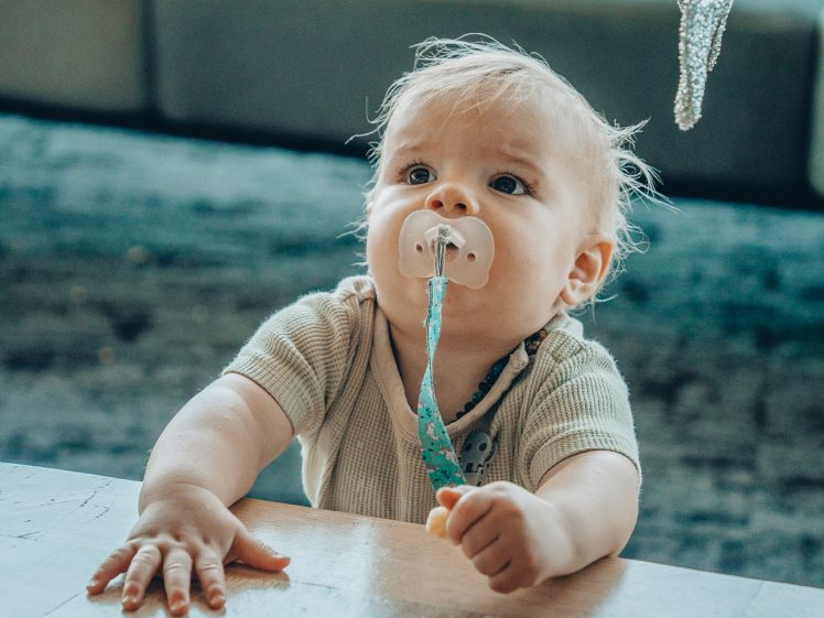 Image of A baby with a dummy in their mouth reaches one hand towards a table and looks up at a grey textured object hanging above them. The background shows a blurred sofa and window.