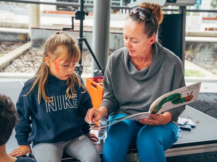 Image of A woman reading a picture book to a young girl with face paint, while a boy sits nearby. They are seated together indoors on a platform, and the girl is focused on the book.