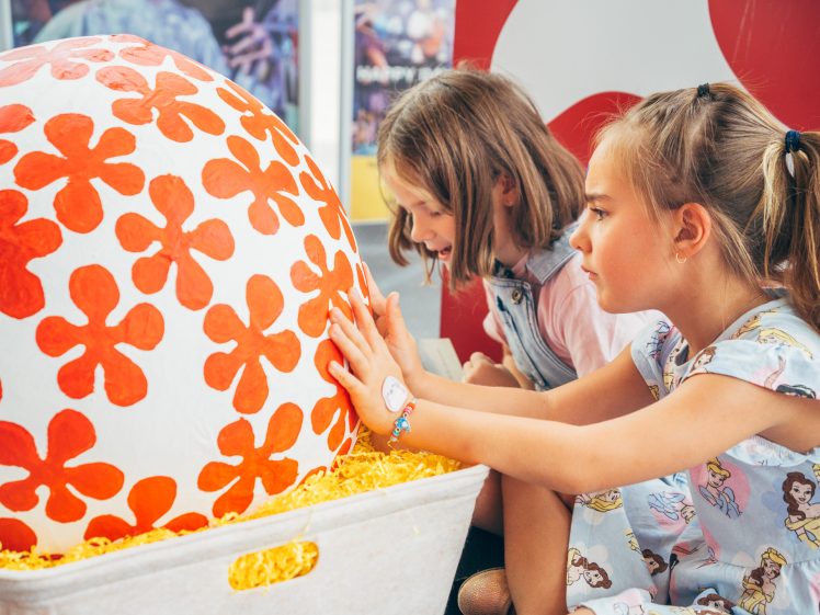 Image of Two young girls closely examine and touch a large white ball decorated with bright red floral patterns, sitting in a bin filled with yellow shredded paper.