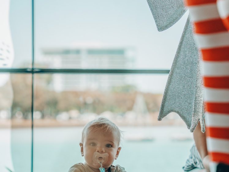 Image of A baby with a dummy looks up while leaning on a table indoors. In the foreground, a person in red and white striped tights and a shiny silver accessory is partially visible. Large windows show a blurred cityscape outside.
