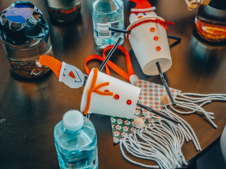 Image of Two handmade snowmen crafts made from white cups, buttons, and orange pipe cleaners lie on a table surrounded by craft supplies, water bottles, and wool.